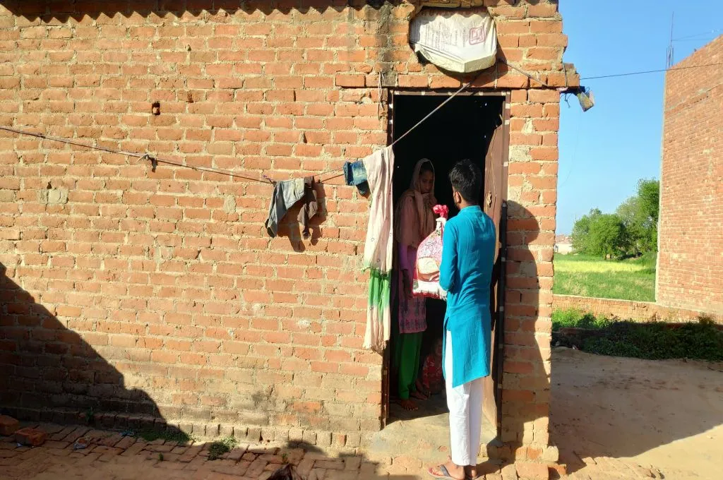 The image shows a young man standing with a woman at the entrance of her house. The house is made of bare bricks with a tin roof. The man is handing the woman a sack of food rations.