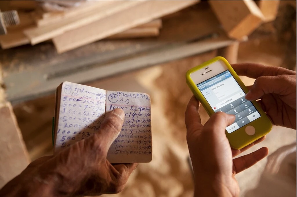 The image is a close-up of two people's hands. An elderly person is holding a small notebook in their hand with rows of numbers written on the page. Next to them, the person is inputting them information on a contacts page open on their mobile phone screen._COVID-19 Jharkhand relief mobile application