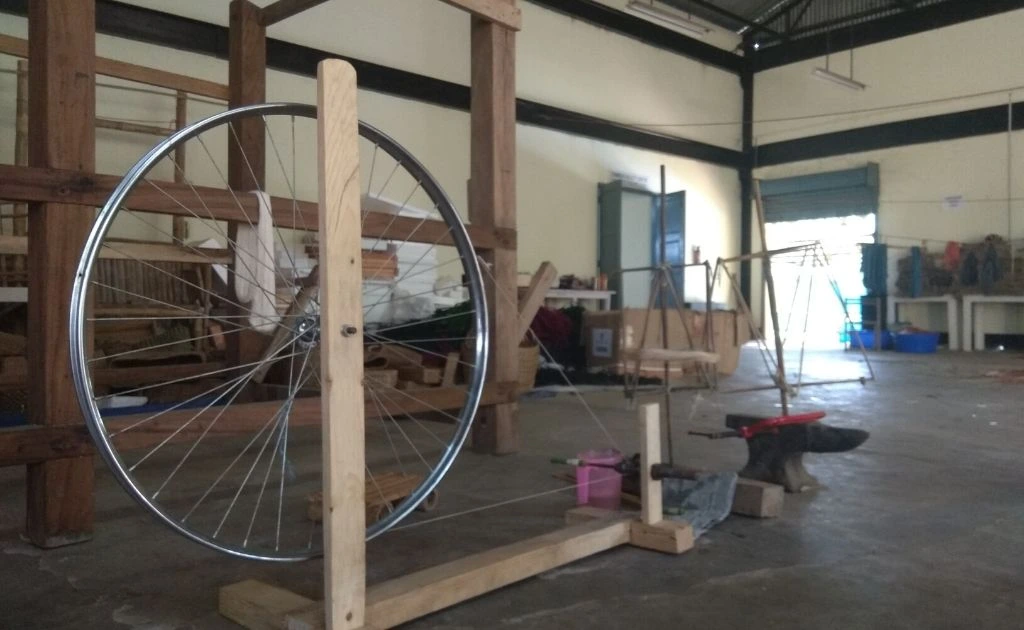 The image shows a large charkha or traditional spinning wheel that is used to create yarn and fibre used in handloom weaving. The charkha has a large metal wheel fixed within a wooden structure.