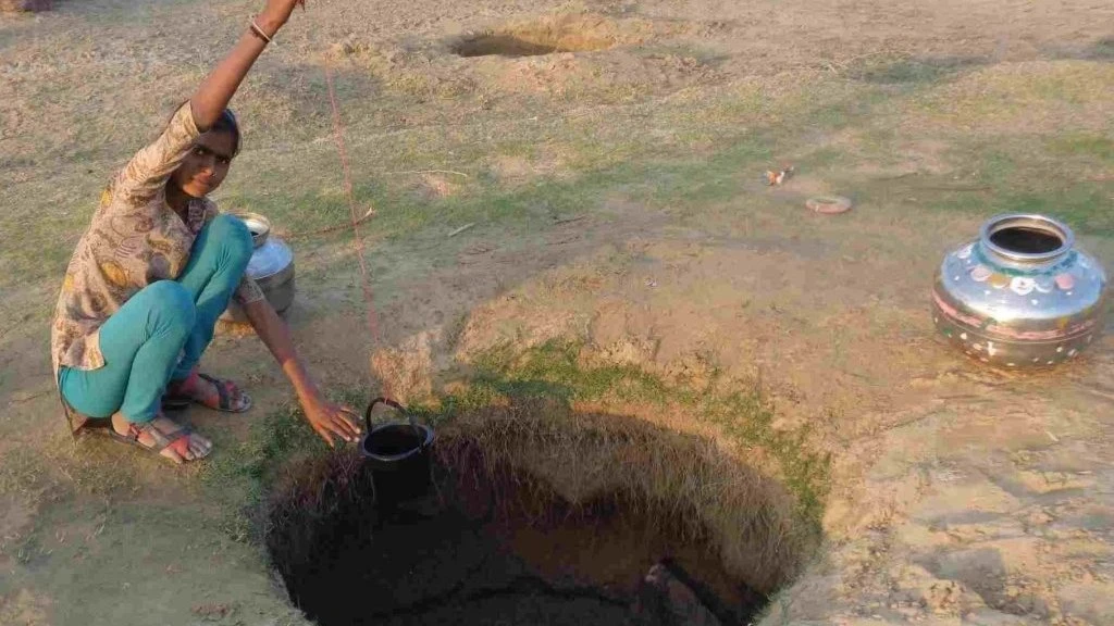 The image shows a young woman drawing water from a small, makeshift dugwell in the ground. There is an aluminium water pot placed next to the well and the woman is pulling a plastic bucket out of the well using a string.