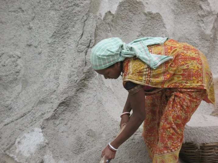 The image shows a woman digging sand using a shovel. The woman appears to be standing in a hole as there is a wall of sand rising behind her.