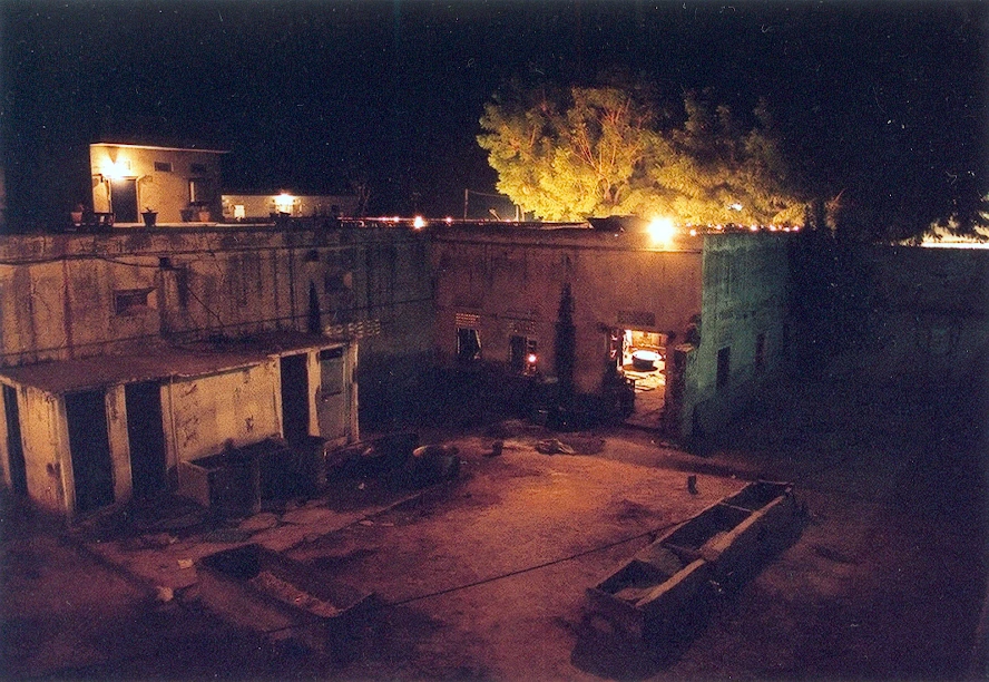 The image shows the outside of single-storey building and its surrounding compound at night-time. The yellow bulb of a streetlight can be seen behind the building and there is a big tree in the background.