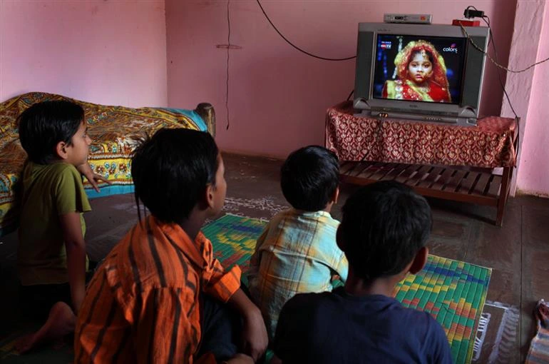 The image shows three children seated on a carpet on the floor watching a programme on the television._Smartphones