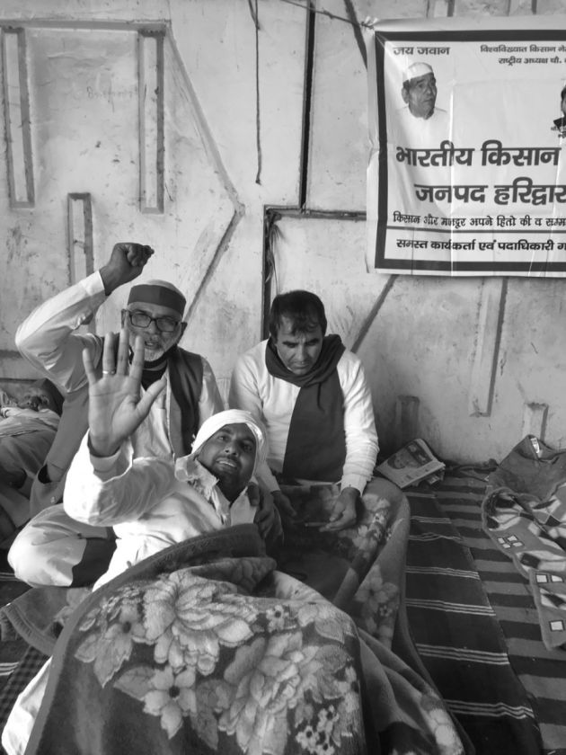 Three men, one with his hands raised, looking into the camera with a banner hanging next to them-farmers protest