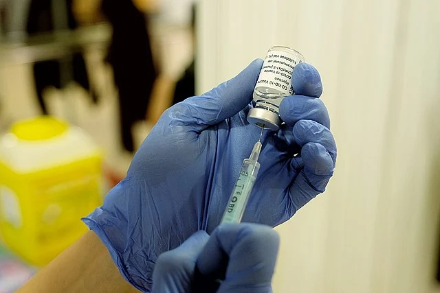 The image is a close-up of a person hands clad in medical gloves. They are filling up a syringe with liquid from a glass vial._Covid-19 vaccine hesitancy