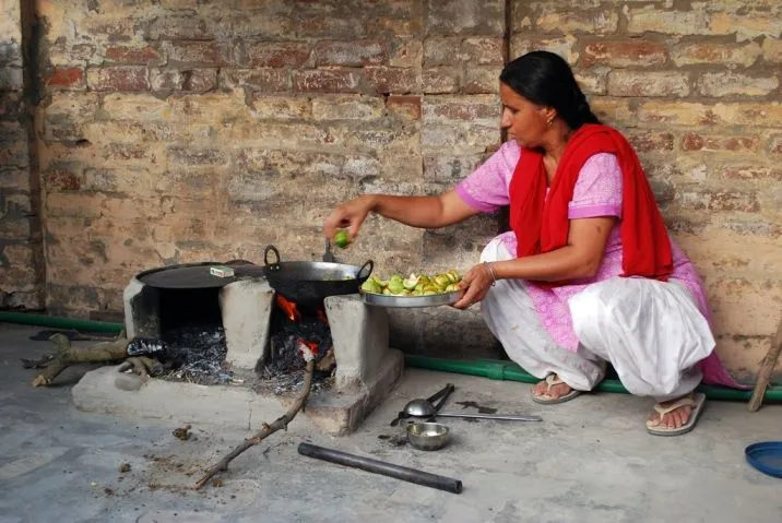 The image shows a woman cooking food over a clay hearth outside her house. There are two cooking vessels placed over a fire and the woman is putting vegetables in the kadhai._COVID-19 gender-based discrimination