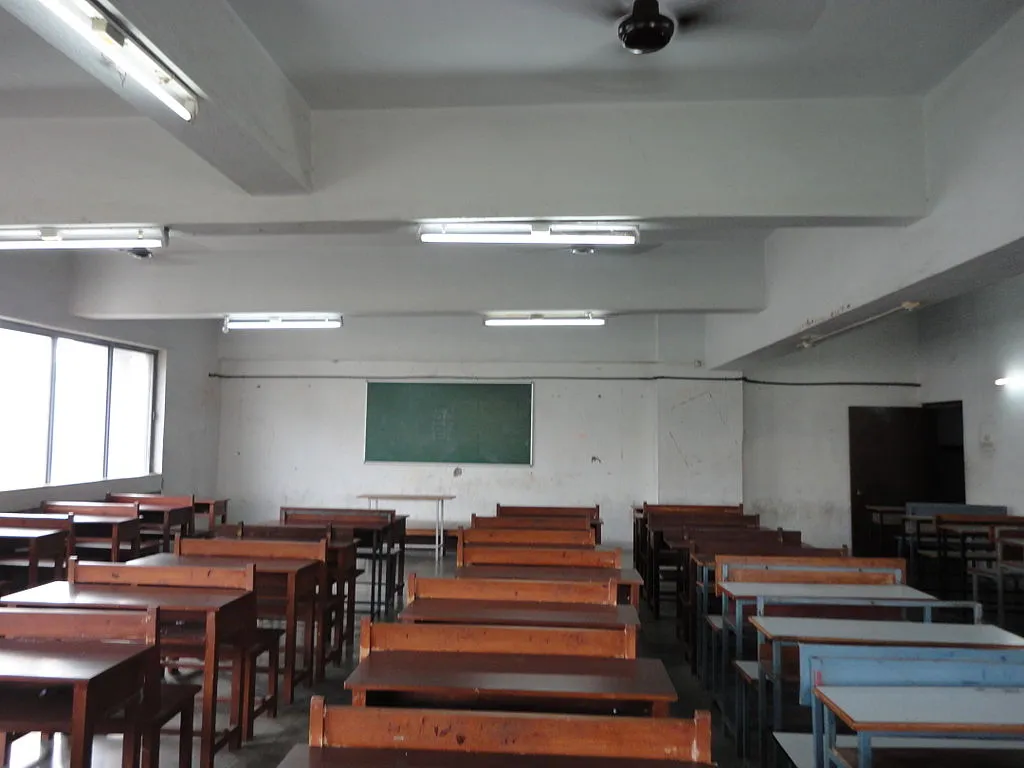 The image shows an empty classroom with rows of wooden benches and desks._COVID-19 vaccination