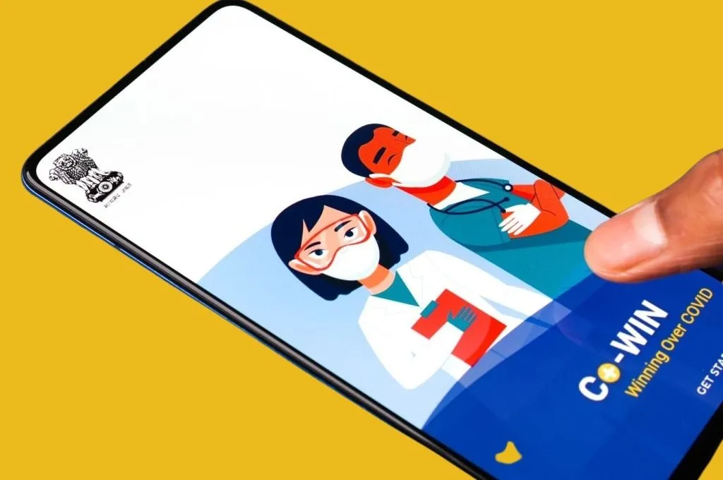 The image is a close-up of a mobile phone screen held in a person's hand. The screen is displaying the landing page of the government's CO-WIN app with an illustration of two doctors wearing face masks._Vaccine hesitancy