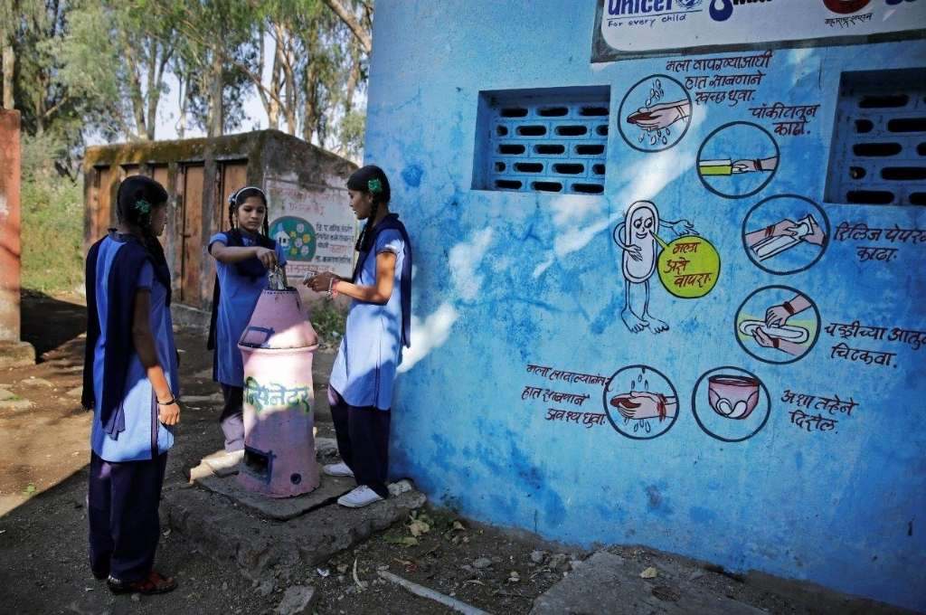 three girls standing around an incinerator as the girl in the middle disposes a menstrual health product into the incinerator. There is a blue wall to their right with wall art about how to safely use a sanitary napkin-menstrual health and hygiene COVID-19