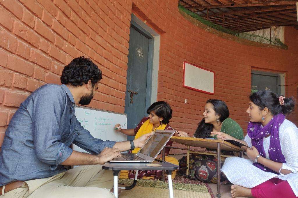 Four people sitting on the ground. From left to right: a man sitting in front of a laptop facing the next woman to his right. A woman writing on a whiteboard with a marker. A woman looking at a whiteboard. Another woman pointing at the whiteboard speaking to the team-education nonprofit