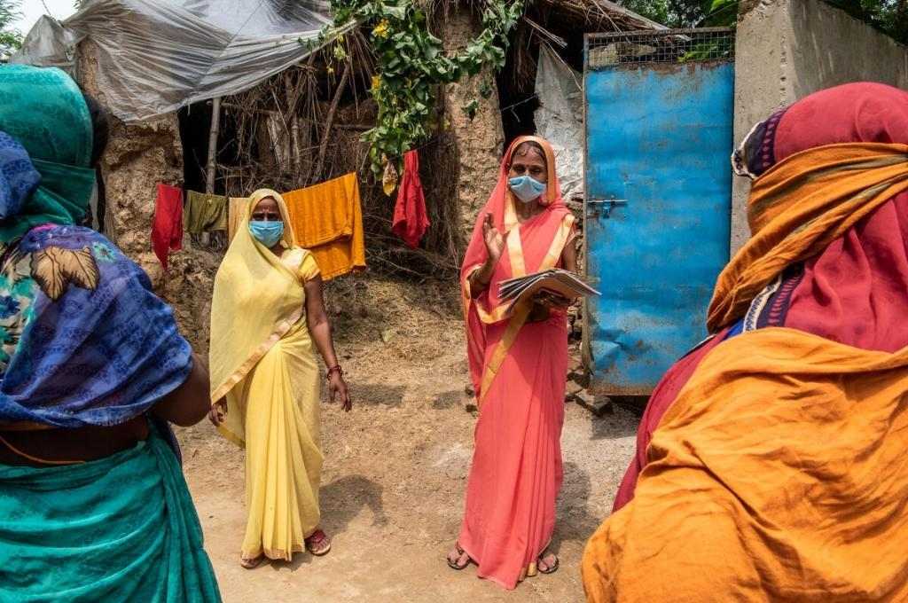 two women facing the camera two women facing the camera, one holding a book, talking to two women whose backs are facing the camera. The two women facing the camera are engaged in going door-to-door to distribute rations-civil society organisations