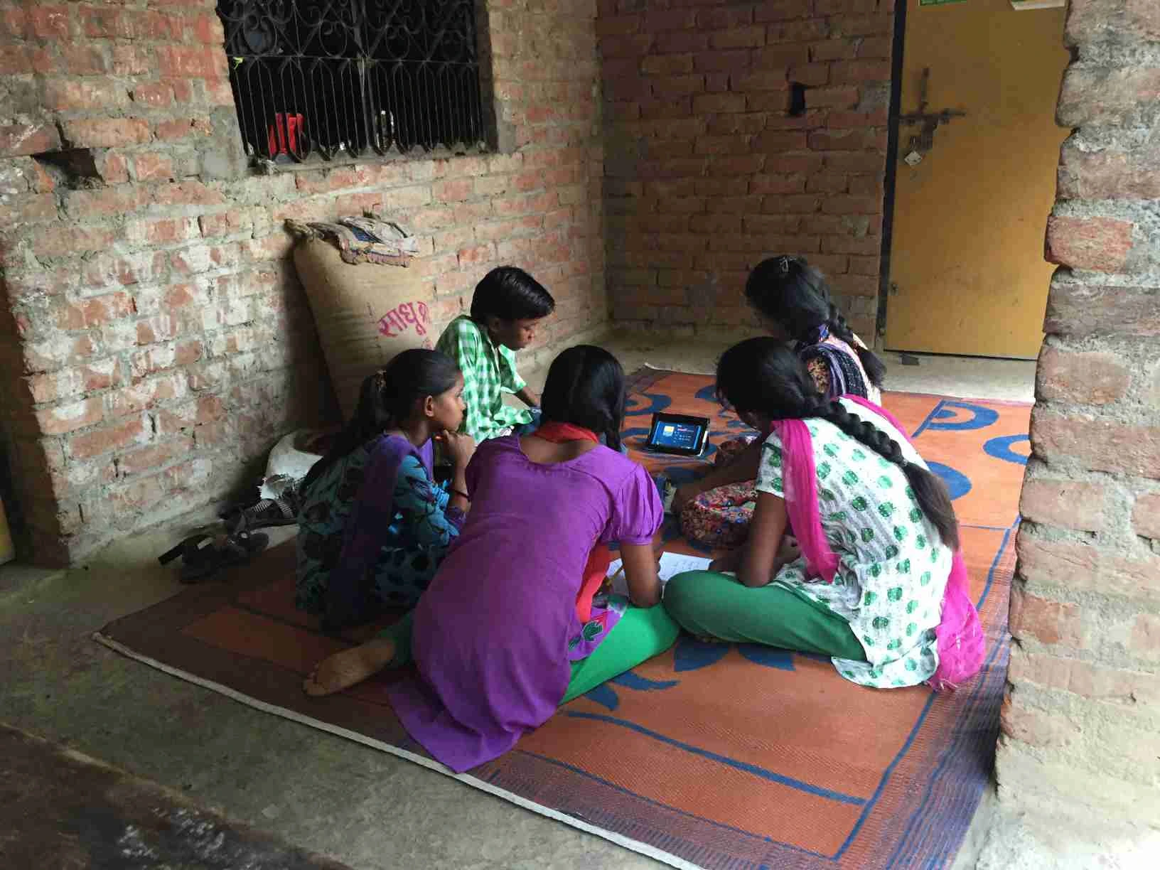 The image shows a group of children sitting in a semi-circle on a carpet on the ground. There are all looking at a phone screen which has information displayed on it. The children also have notebooks open in front of them._E-learning