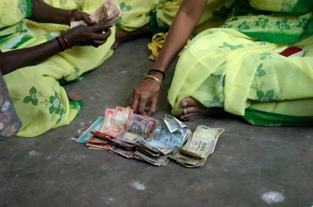 The images shows small stacks of rupee notes arranged according to their value and placed on the ground. Two women sitting on the ground can be seen partially, and one woman is counting a separate stack of notes held in her hands._Loan moratorium second wave