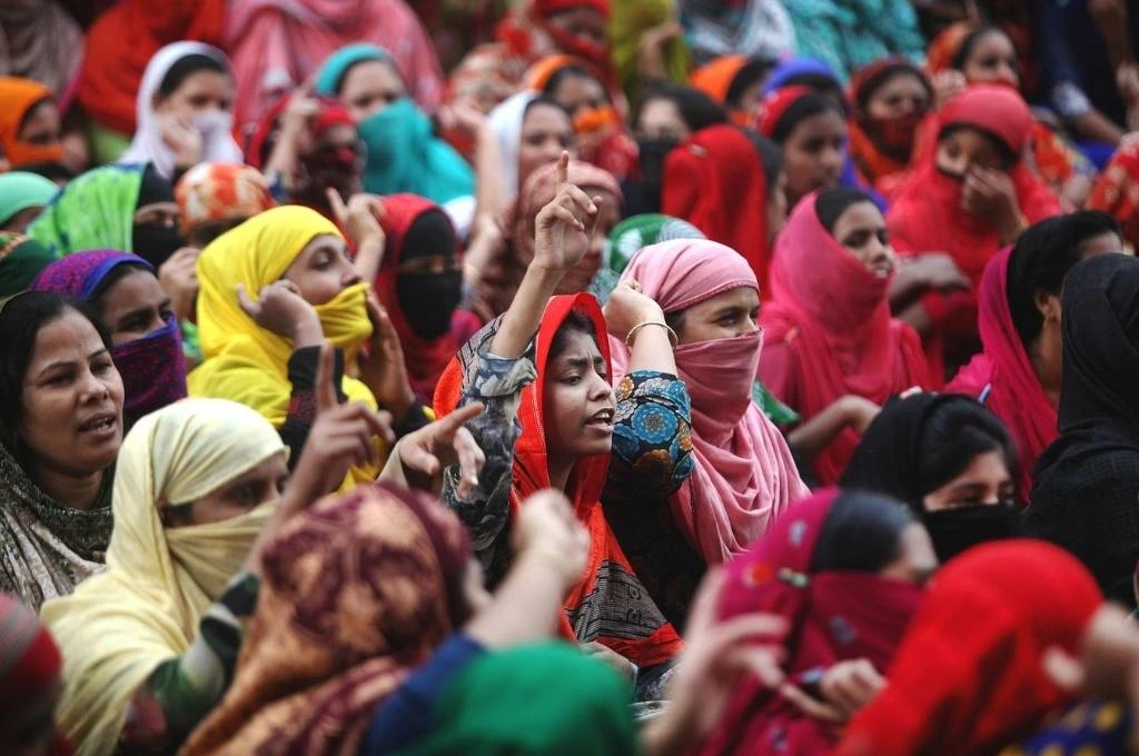 The image shows a group of women sitting together, many with their faces or heads covered with dupattas. A young woman is speaking with her arm extended and a finger raised in the air._COVID-19 awareness