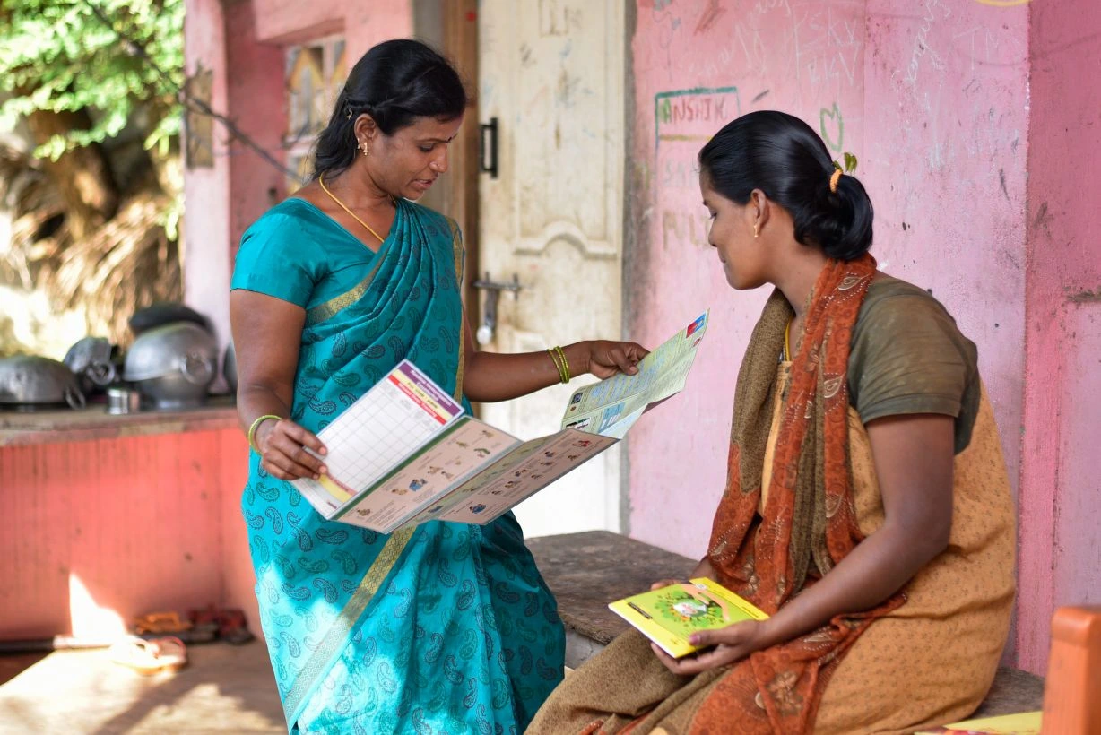The image features two women in the courtyard of a house. One of the women is wearing a sari and she is reading out loud from a pamphlet that she is holding in her hands. Beside her, a pregnant woman is seated on a wooden bench and she is also looking at the pamphlet._ASHA workers