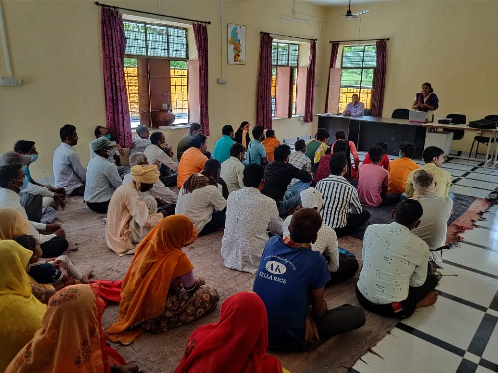 The image shows a group of men and women sitting in rows on the ground in a room. They are listening to a woman who is standing behind a table in front of the room. There is a laptop open in front of her and a man is sitting next to her._COVID 19 vaccine