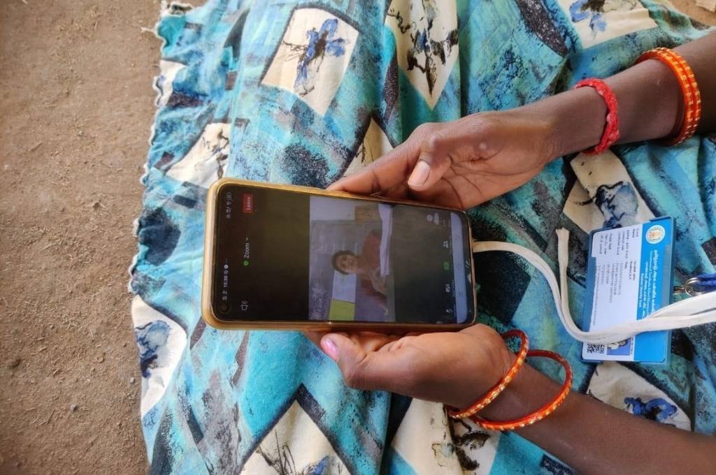 An image of a woman holding a mobile phone with a zoom video playing on it. We can see her saree, there are bangles on her hand, and we can see her id card hanging. As we move from digital learning to physically attending schools again, what steps can policymakers take to create a supportive and enabling environment for government school teachers-teachers