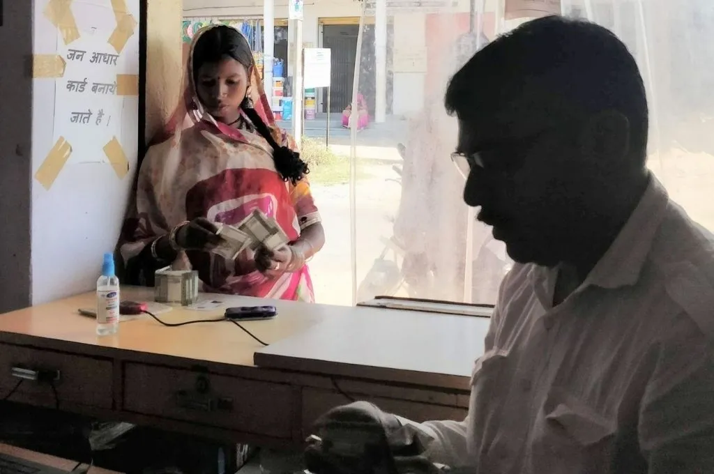 The image shows a young woman standing at the counter of a small shop in a rural area. She is counting money. The sign on the wall next to her reads 