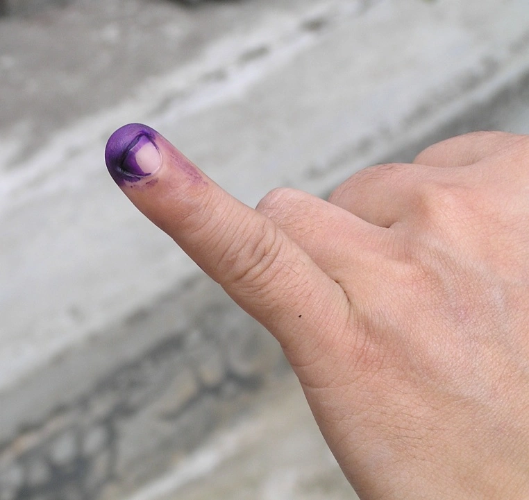 The image shows a person's hand with their smallest finger extended. The top part of the finger has been dyed with blue ink._NRC Assam