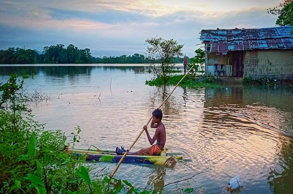 The image shows a young child rowing a narrow wooden boat through water. There are trees and bushes growing around the edges. Behind the child, there is a house made of tin sheets that is surrounded by the water._Assam flood