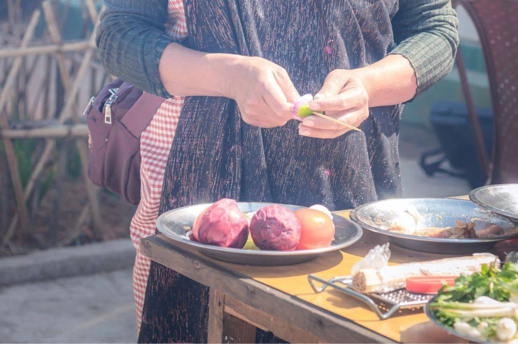 The image is a close-up of a woman's hands as she uses a knife to chop vegetables. In front of here, there is a table on which there is a plate of onions and tomatoes. There are other utensils and vegetables on the table._Indigenous food