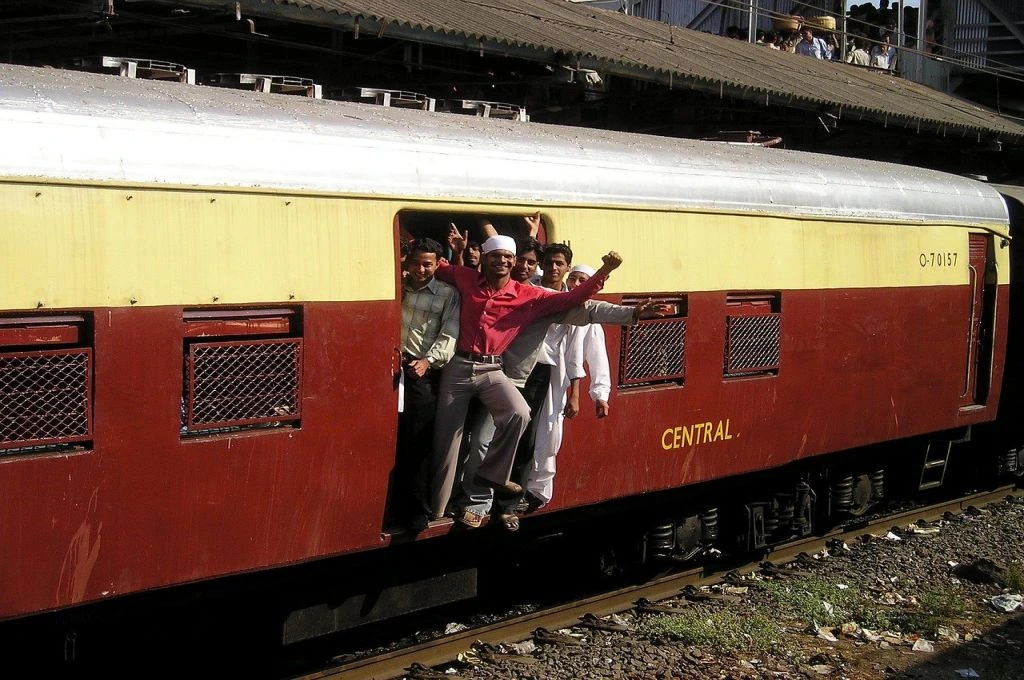 The image features a group of men leaning out of the door of a passenger train. Some of the men are smiling and have their arms raised in the air._Gender