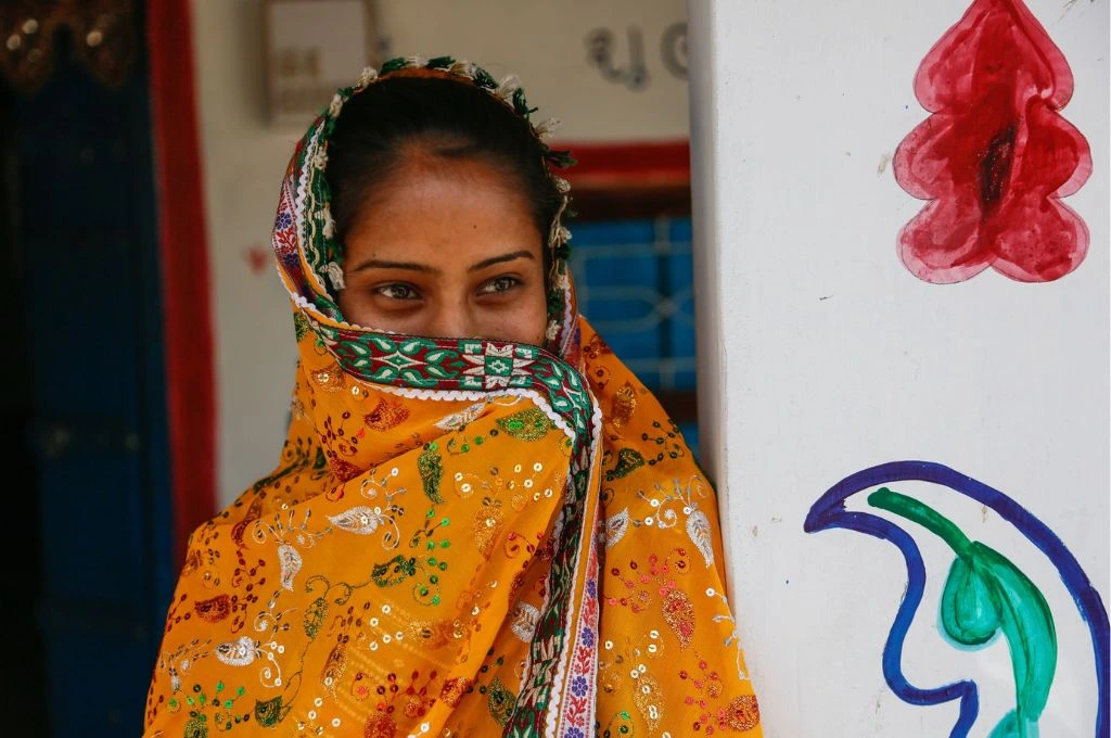 The image features a young woman leaning against a wall that has designs painted on it. The young women has covered the lower half of her face with a dupatta and she is smiling slightly._Migrant