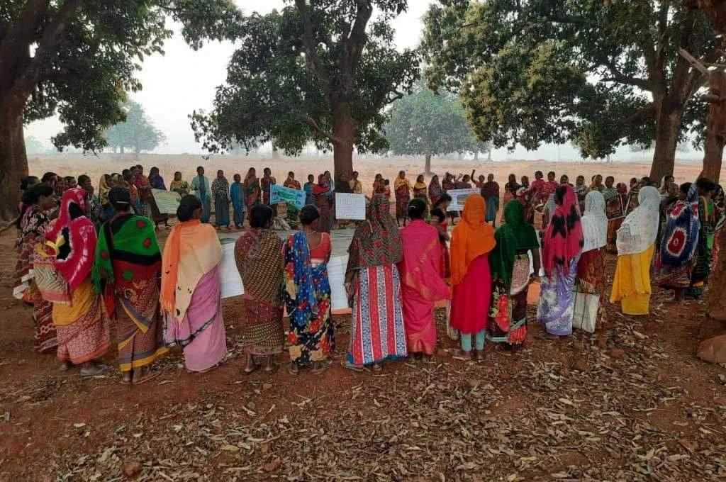 The image features a large group of women standing in a circle. They are surrounded by trees. Some women are holding posters in their hands. Behind them, a large open ground with trees stretches into the distance._Forest fires
