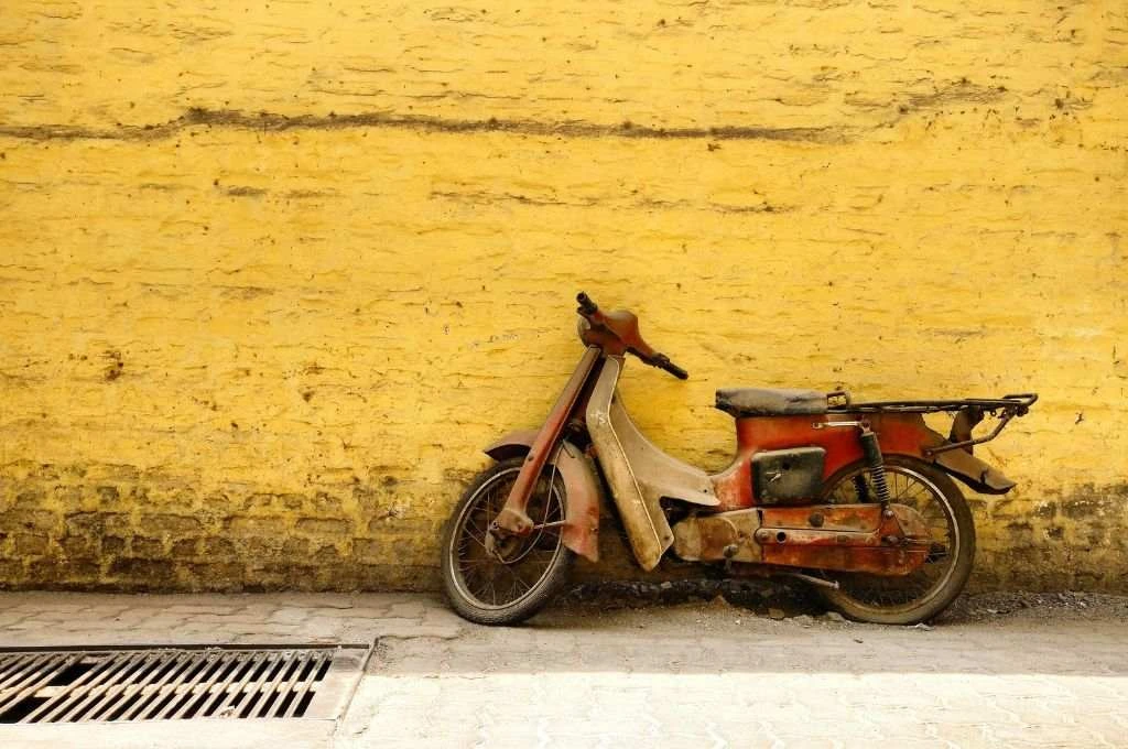 The image shoes an old red-coloured scooter parked against a yellow brick wall._social worker
