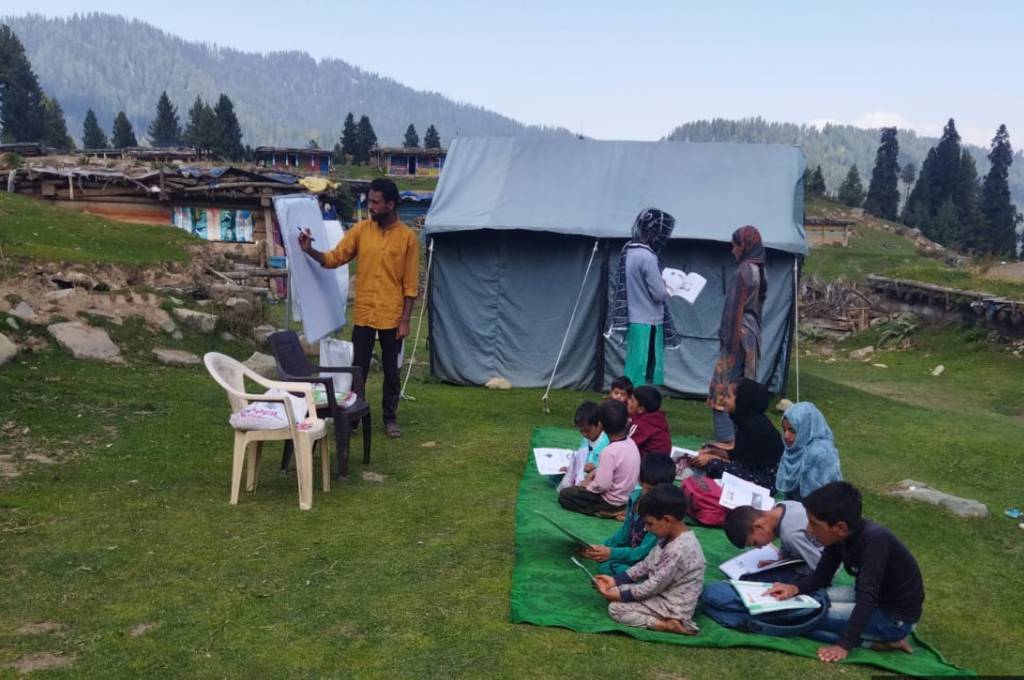 A teacher teaching students in the field in Kashmir valley. The students are sitting on the ground on a mat. There is a tent in the background_Pastoralists in Kashmir