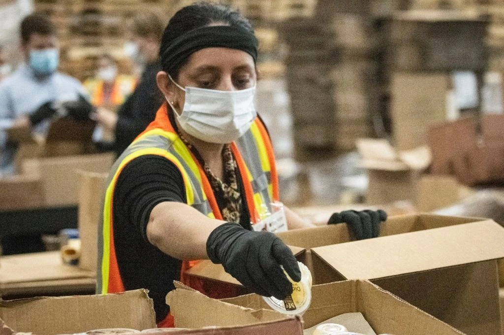 A woman wearing a mask, an orange safety vest, and gloves packs things in a box in a warehouse._women workers