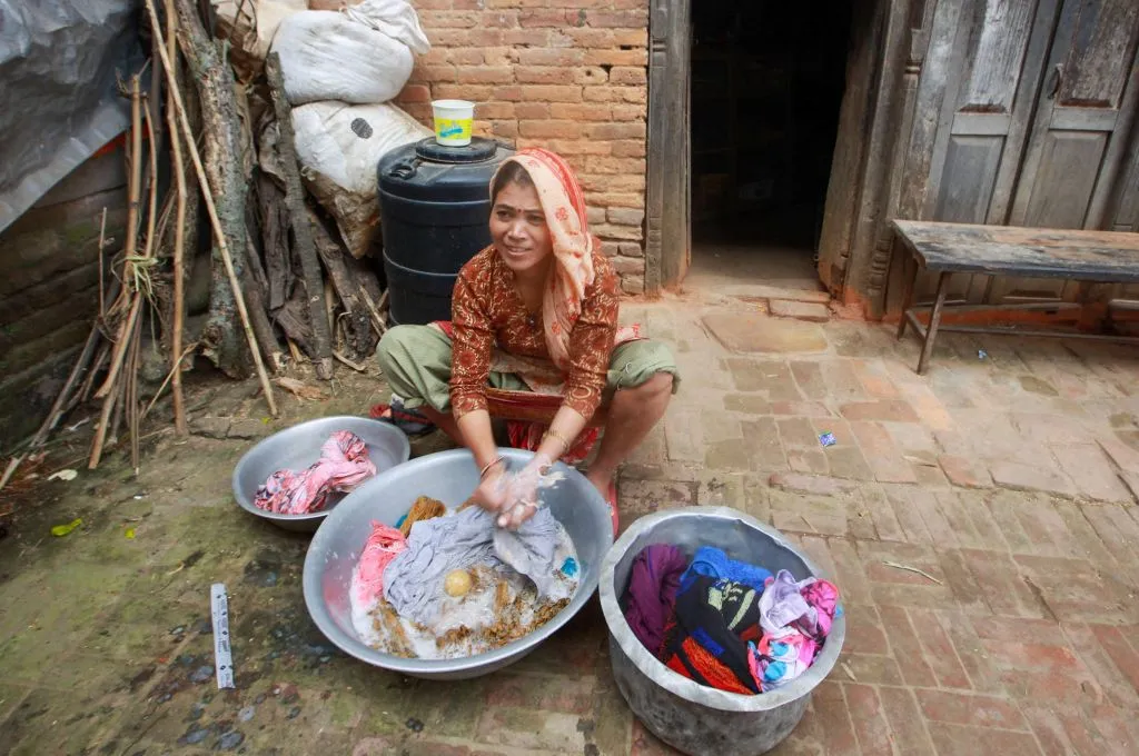 The image features a woman washing clothes by hand. The clothes are being washed in a big metal utensil and there are two separate utensils on either side containing clothes._hawker