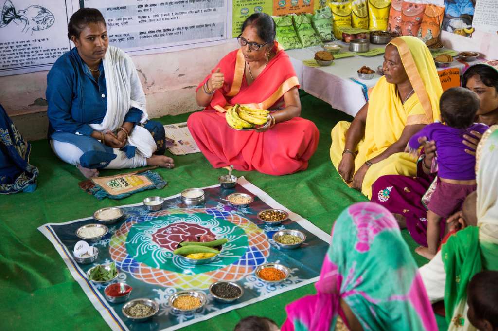 A gathering of women at an Anganwadi centre on the benefits of eating nutrtious foods with a display of various pulses, grains, and fruits_non-communicable diseases