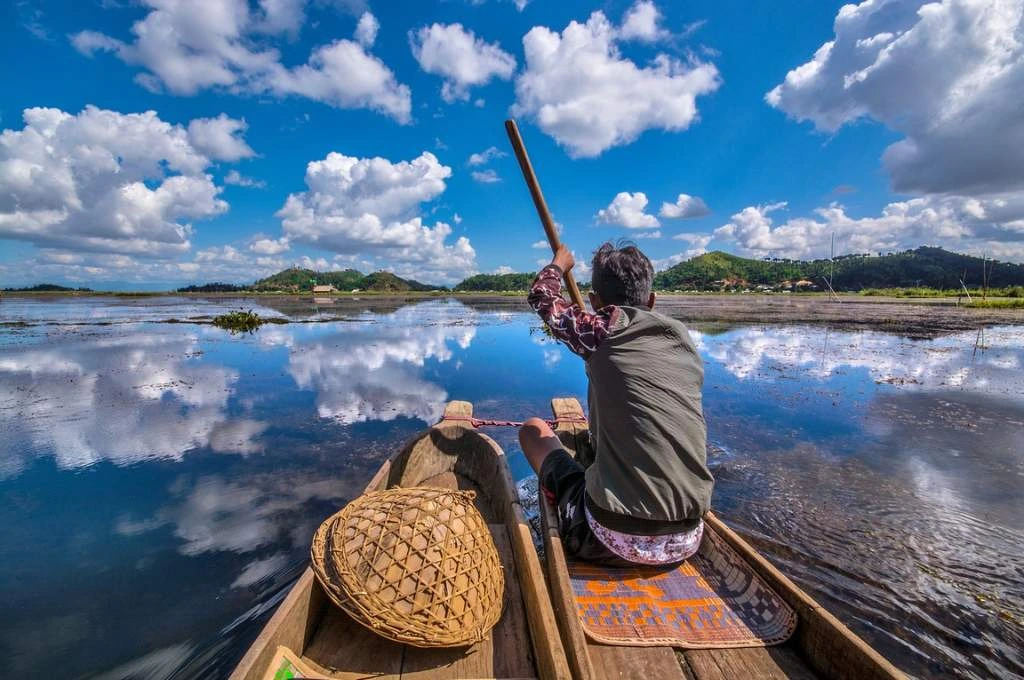 The image shows an old man rowing two narrow wooden boats that are tied together in a vast body of water. There are small, green hills in the distance._Manipur violence