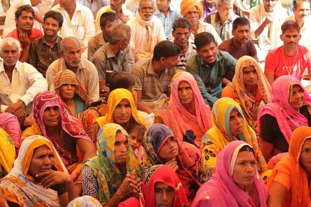 The image features a group of men and women sitting in rows on the ground. They are of varying ages and are presumably in a village._Microfinance institutions