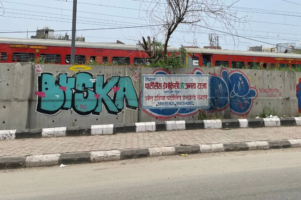 The image is of a bicycle path on a road attached to which is a concrete boundary wall with grafitti and painted signs on it. Behind the wall, a train passes._Sawda Gherva Village