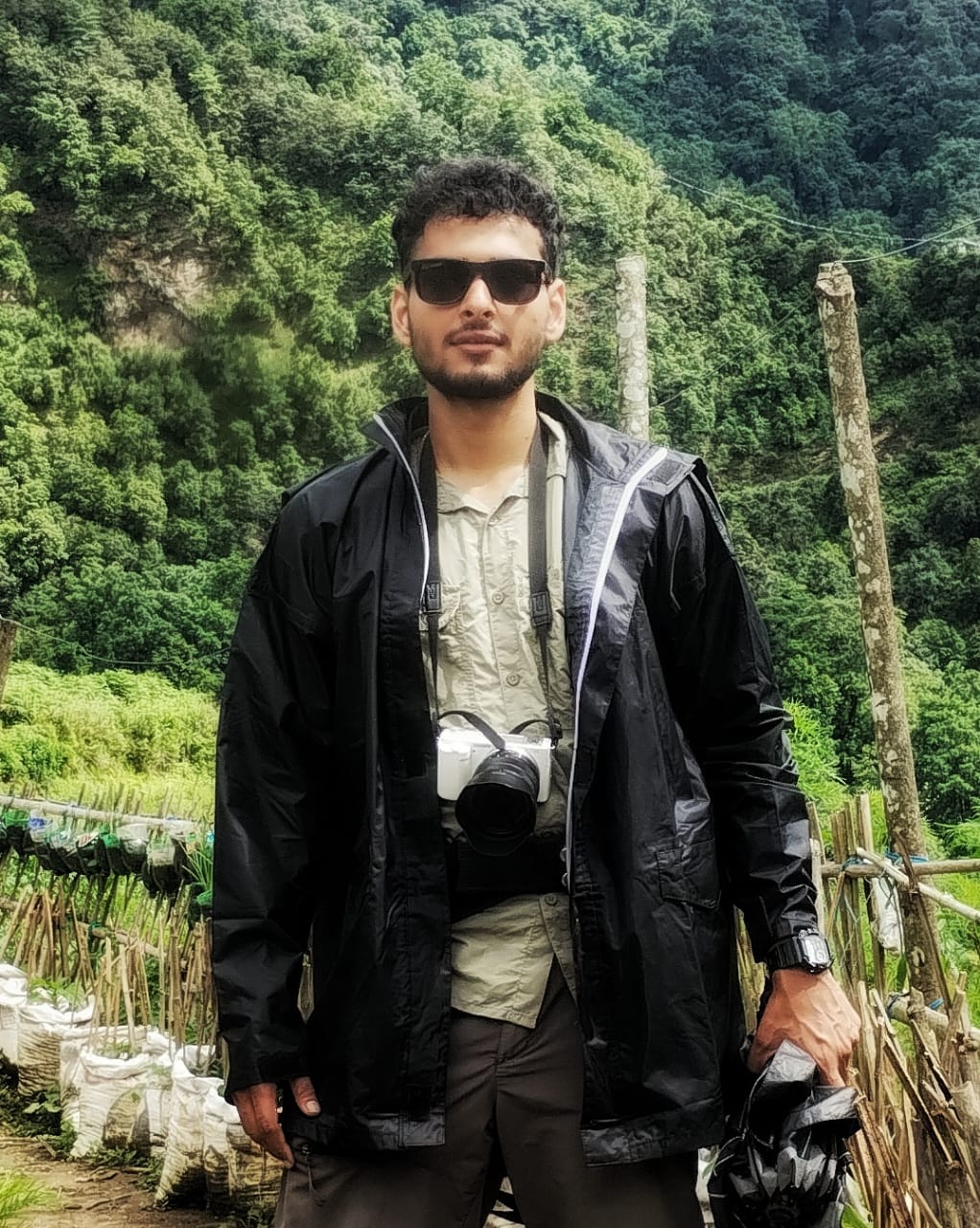 A man wearing a black jacket and sunglasses. He has a bamboo pool and greenery as his backdrop