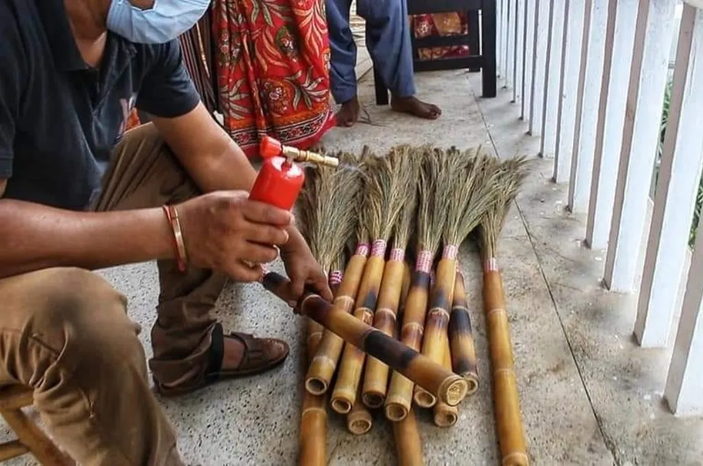 A person uses a small metal can put black paint on a bamboo stem to make brooms while a bundle of finished brooms made of bamboo stems and grass lay on the ground- Bamboo products