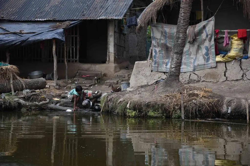 A child washes utensils on the bank of a water body with a house made of mud with a thatched roof and another house made of cement with a tin roof in the background._children and climate