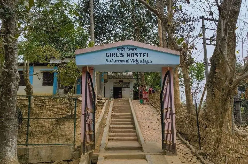 The image shows the entry gate of the Girl's Hostel of Ratanmani Vidyalaya. Behind the gate, there are stairs leading up to a building with two young girls standing outside._Language barriers in education