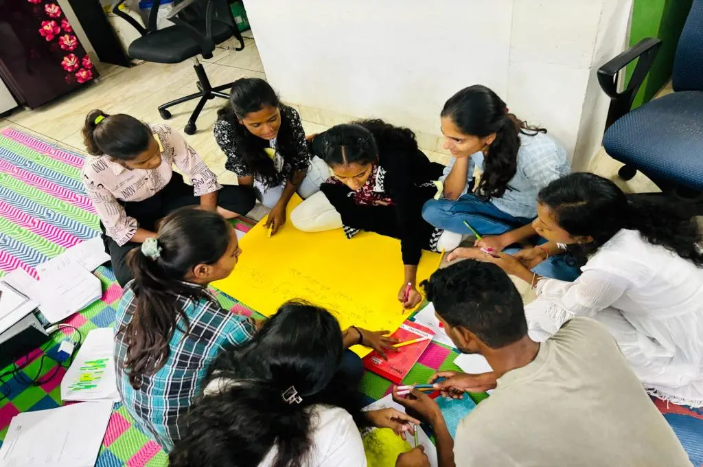 The image features a group of adolescents and young adults sitting together in a circle and holding a discussion. A young woman uses a sketch pen to write on a yellow chart paper that is placed in the middle of the circle._Care leavers