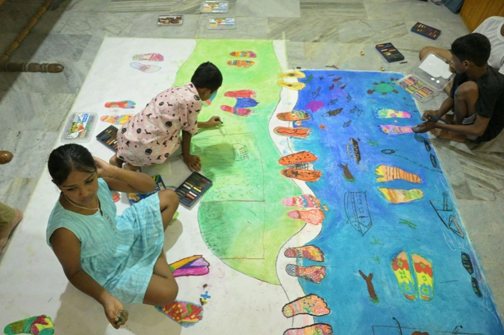 The image shows three children drawing and colouring in a map of Tezpur and Brahmaputra. The children have drawn colourful footsteps along the bank of the river and in the water._Memory mapping
