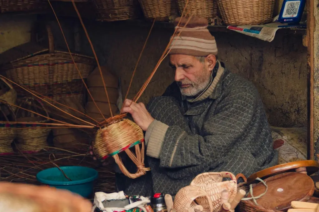 The image features an old man sitting in a shop and weaving a cane basket by hand. There are woven baskets of various sizes and other goods in the background._Kashmiri artists