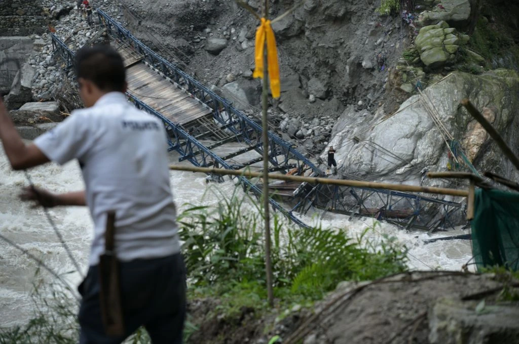 The image shows the collapsed Sankalang Bailey Bridge. Parts of the bridge are broken down and submerged in the Teesta river as water flows by in high speed. There are two people on either side of the collapsed bridge._Memory mapping
