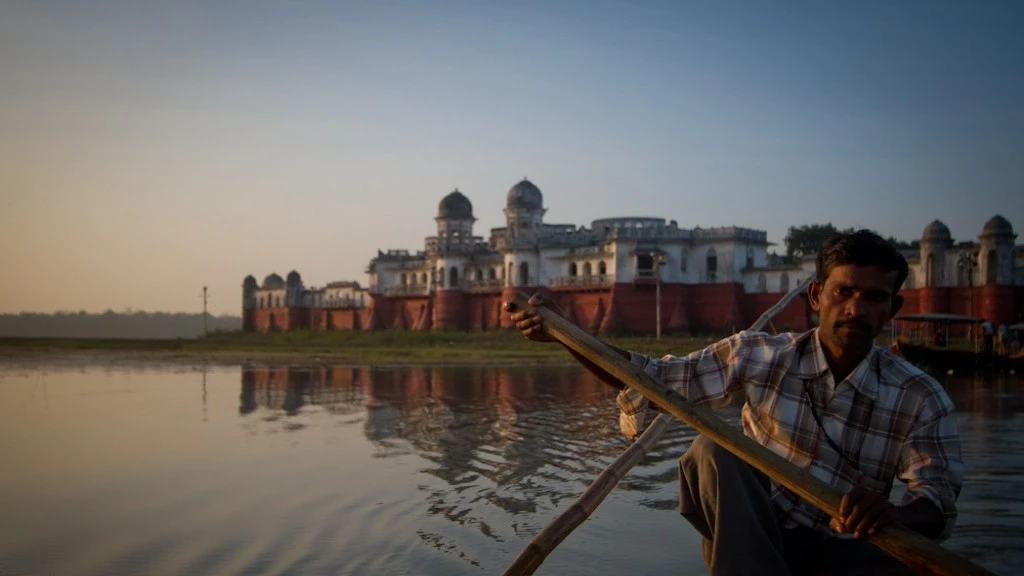 The image features a man rowing a wooden boat. Behind him, on the edge the water, is an old palace._Educational inequity