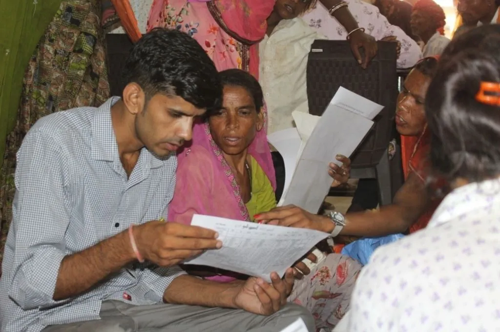 a man and a woman go over a document together, with several other people in the background and around them--Pradhan Mantri Suraksha Bima Yojana claim