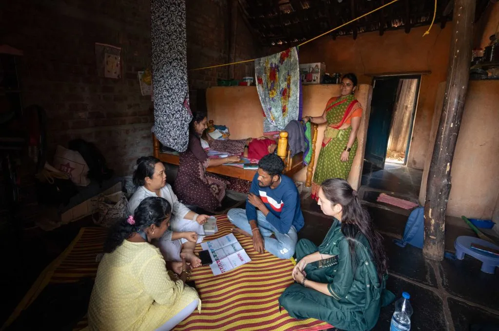 The image is of the inside of a person's house. In the background, a women lies on the bed as a another women sits on the floor beside her and talks to her. Another women stands by the bed. In front of them, three women sit and talk with a young man._Maternal and newborn care