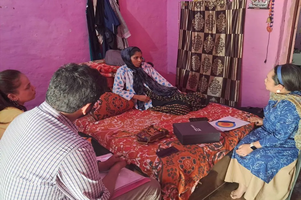 A woman sits on the bed. There are three other people sitting around the bed and the group is in the middle of a conversation._Maternal and newborn care