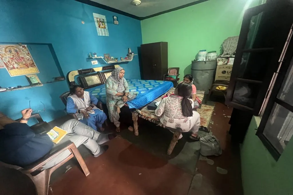 The image features the inside of a person's home. A women sits on the bed. There are four other people in the room, two of whom are young women sitting at the foot of the bed. The women are having a conversation._Maternal and newborn care