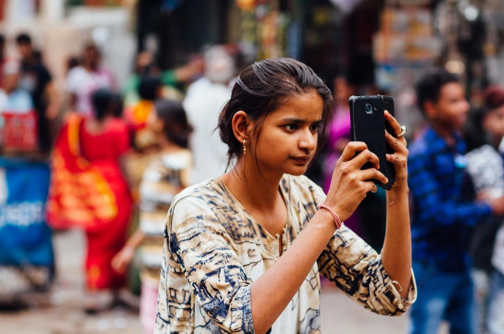 A young woman in a crowded public space takes photos using her mobile phone._Caste and digital divide